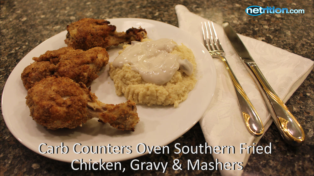 A plate of crispy oven-fried chicken drumsticks is served alongside creamy mashed potatoes topped with gravy, with a fork and knife resting on a folded napkin on a granite countertop.