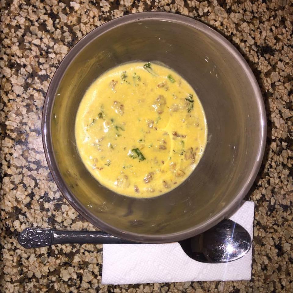 A silver bowl contains a creamy mixture with bits of meat and green herbs, resting on a speckled countertop next to a folded napkin and a shiny spoon.