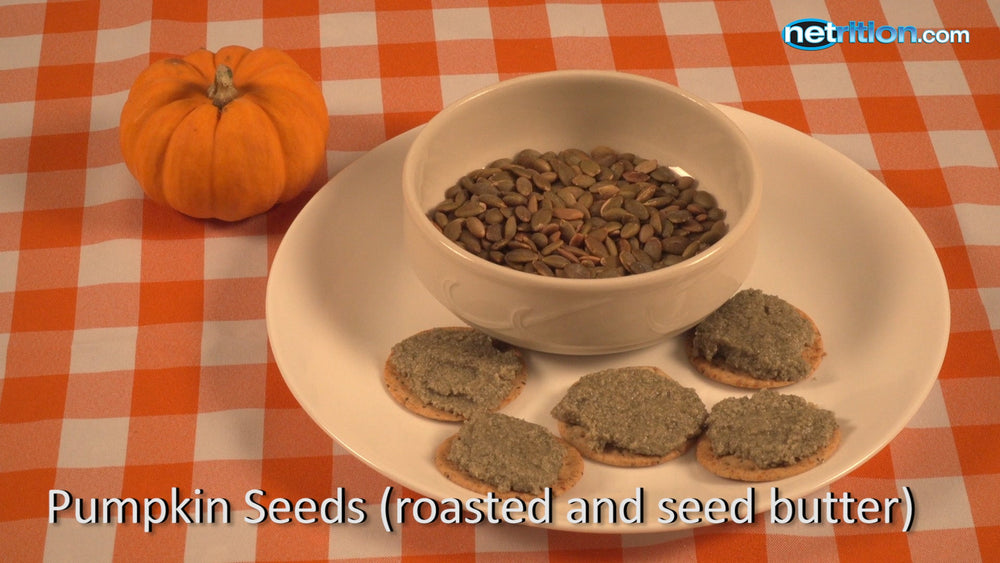 A small orange pumpkin sits beside a white plate with three crackers topped with a gray seed butter, and a bowl filled with green roasted pumpkin seeds, all on an orange and white checkered tablecloth.