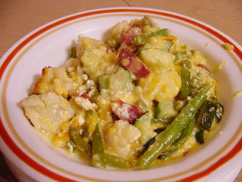 A bowl of mixed vegetables, cheese, and bacon is served, showcasing a colorful blend of green beans, zucchini, and creamy textures, set against a light wooden surface.