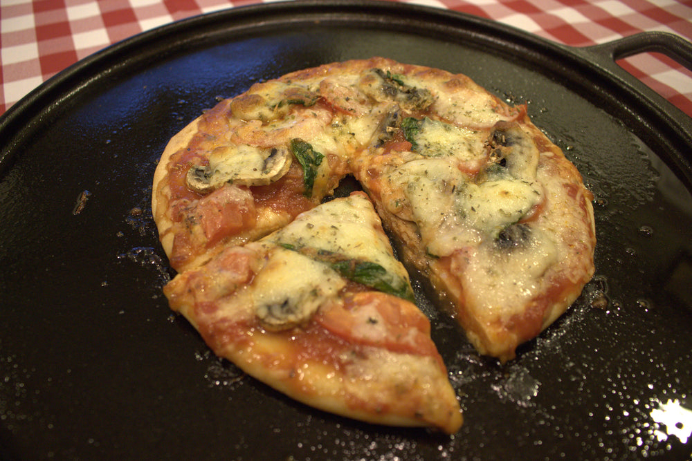 A freshly baked pizza topped with cheese, mushrooms, and spinach sits on a black serving tray, with one slice cut out, on a red and white checkered tablecloth.