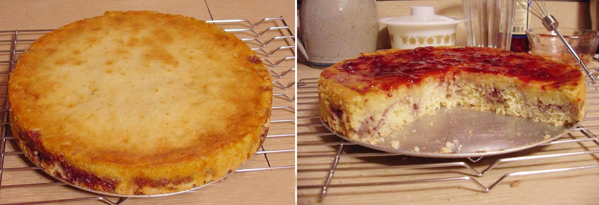 A round cake with a golden-brown crust sits on a wire rack, with a slice removed revealing a moist interior and a layer of red fruit jam on top, set against a kitchen background with utensils.