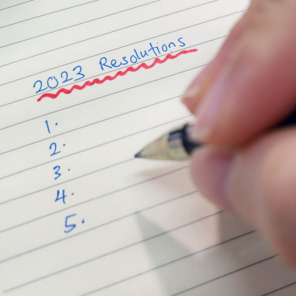 A hand holds a pen poised to write on a lined notebook, with the title "2023 Resolutions" written above a numbered list of five blank lines.