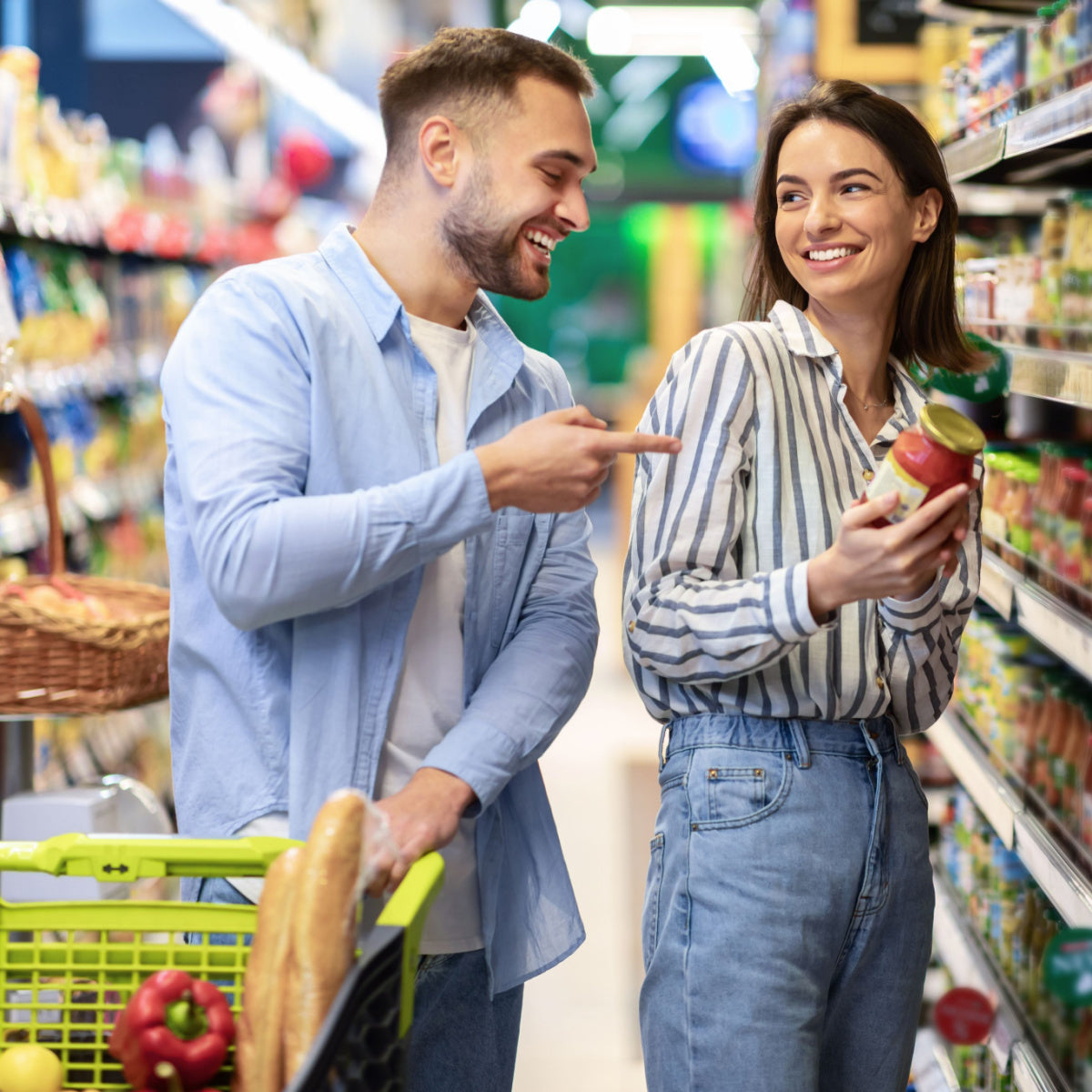 A man smiles and points at a woman holding a jar of sauce while shopping together in a grocery store aisle filled with various food products.