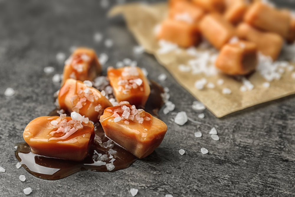 Soft caramel pieces are scattered on a dark surface, some are glistening with a syrupy sheen and topped with coarse salt, while a few more pieces are placed on a crumpled brown paper in the background.