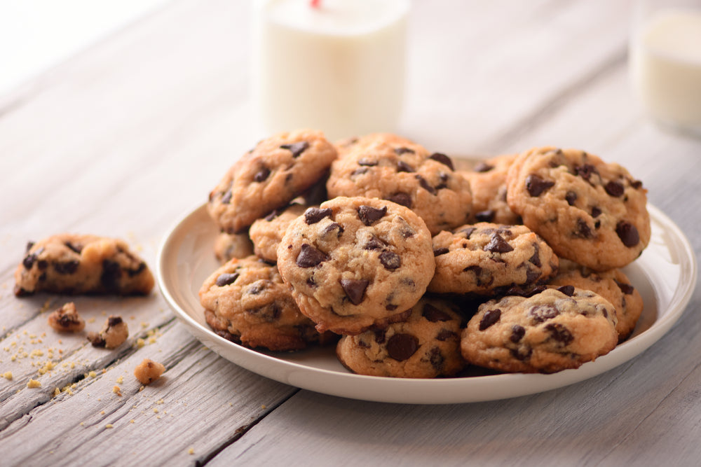 A plate of freshly baked low carb chocolate chip cookies sits on a light wooden table, with a few cookies scattered nearby and a glass of milk in the background.