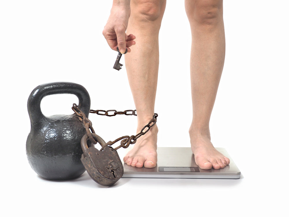 A person stands barefoot on a scale, holding a key while a heavy kettlebell is chained to their ankle, symbolizing the struggle with weight and freedom.