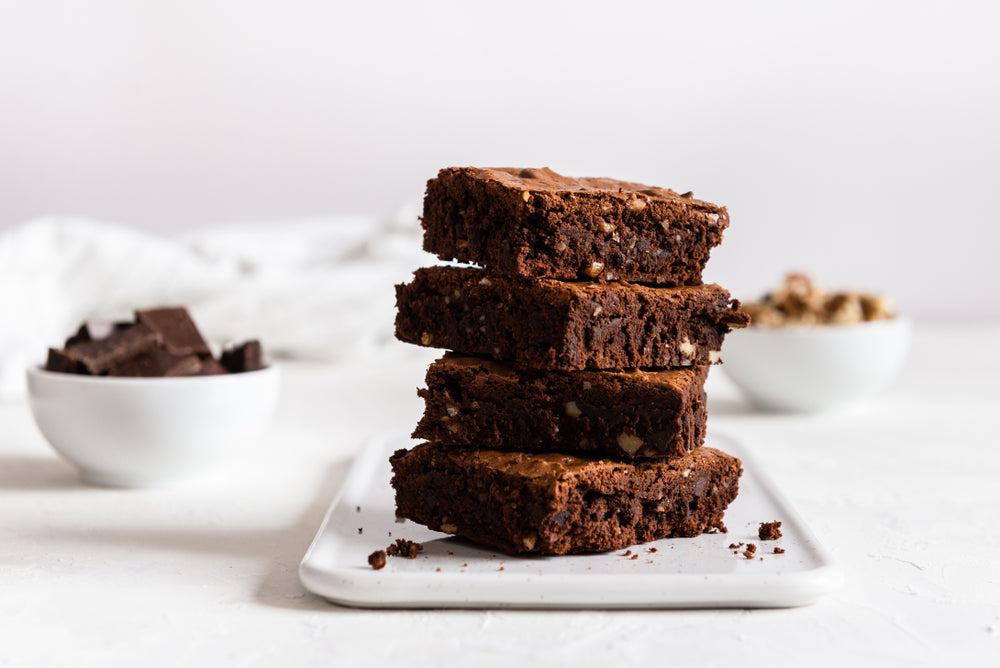 Four stacked brownies with a shiny top sit on a white plate, surrounded by bowls of chocolate chunks and nuts, against a light background.