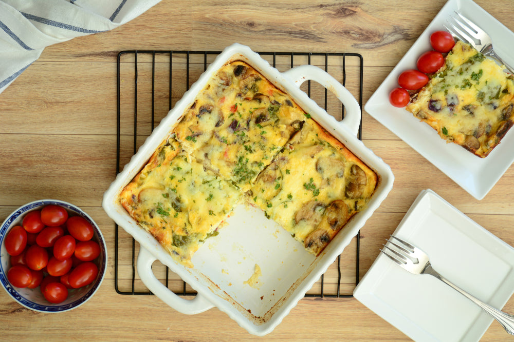 A baked frittata with mushrooms and herbs sits on a cooling rack, alongside a bowl of cherry tomatoes and a plate with a serving of frittata garnished with parsley.