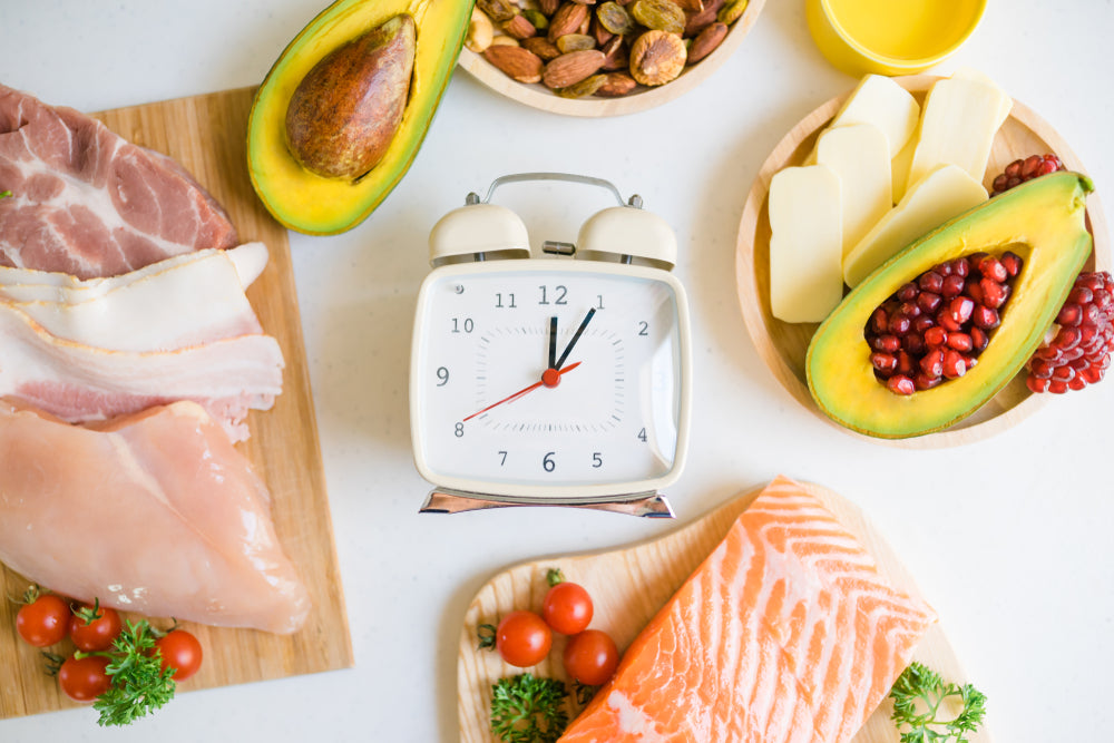 A vintage alarm clock sits among various keto-friendly foods, including avocado, meats, nuts, and cheese, arranged on wooden boards, suggesting a meal prep for intermittent fasting.