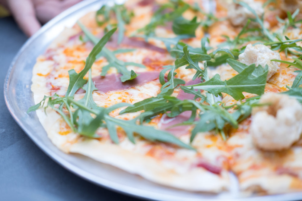 A close-up of a pizza on a silver tray topped with arugula, prosciutto, and small pieces of crispy pork, with a hand partially visible in the background.