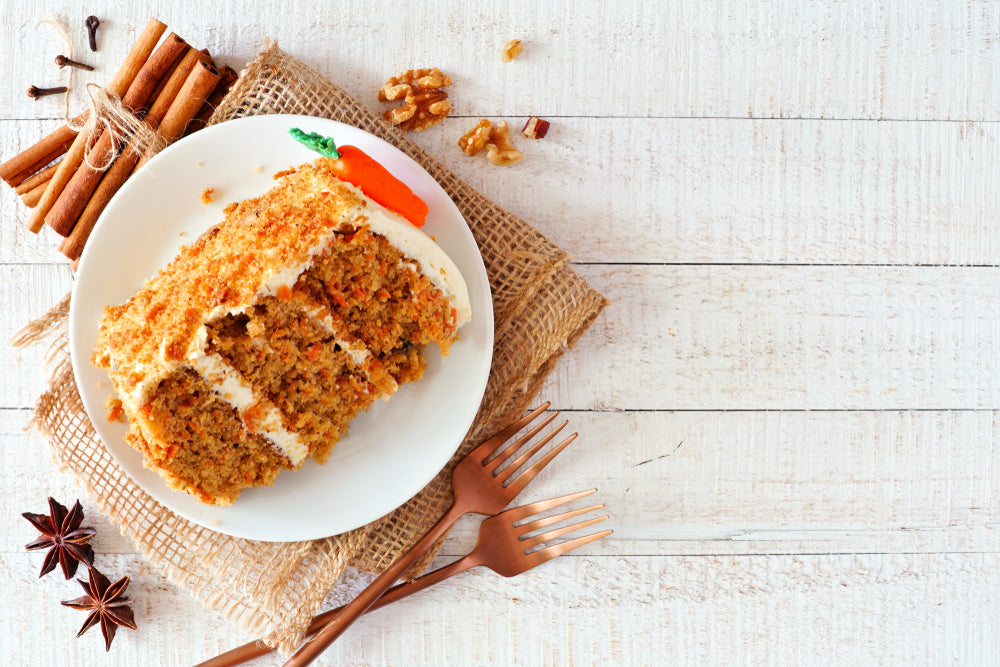 A slice of carrot cake topped with cream cheese frosting is served on a white plate, surrounded by cinnamon sticks, walnuts, and two wooden forks on a rustic wooden table.