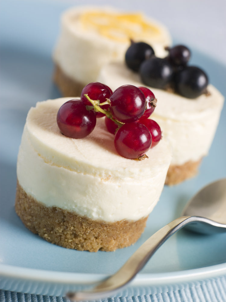 Three small cheesecake desserts sit on a light blue plate, topped with red currants and blackberries, with a silver spoon resting nearby.