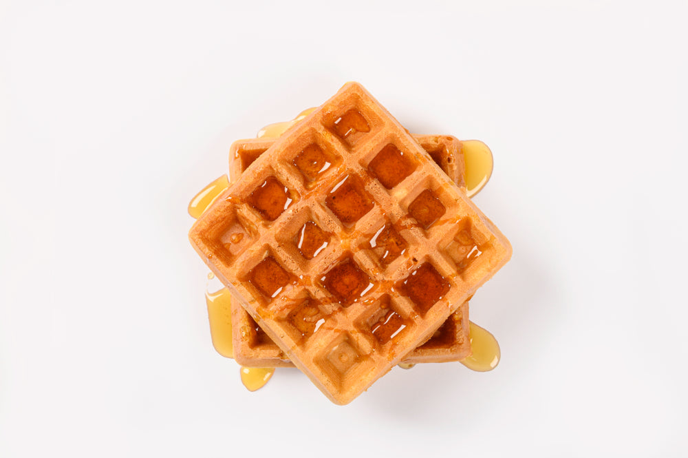 Two golden-brown waffles are stacked on top of each other, drizzled with syrup, and set against a plain white background.