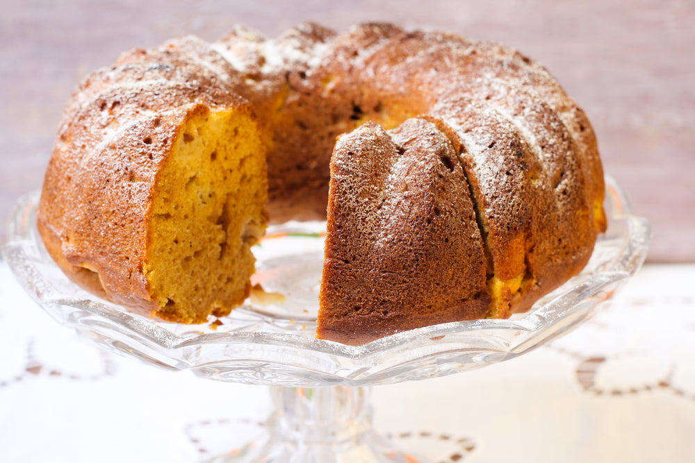 A round, golden-brown bundt cake with a slice cut out sits on a clear glass cake stand, dusted with powdered sugar against a soft, neutral background.