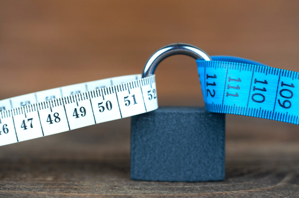 A black padlock sits on a wooden surface, with a white measuring tape and a blue measuring tape looping around its shackle, indicating a theme of measurement and security.