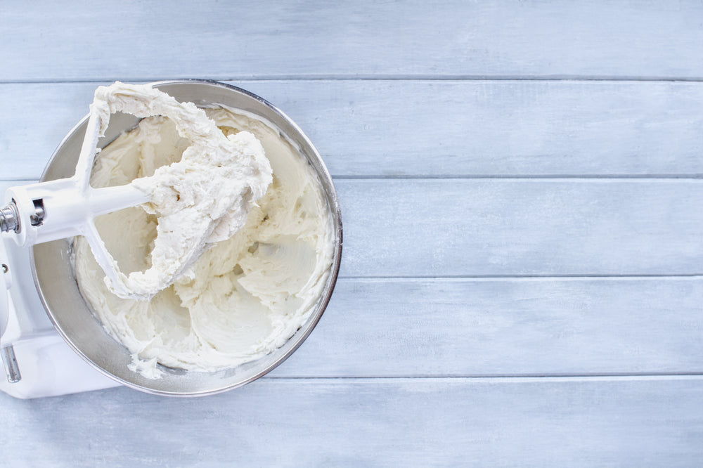 A mixing bowl contains creamy frosting, with a mixer attachment resting on the edge, set against a light blue wooden surface.