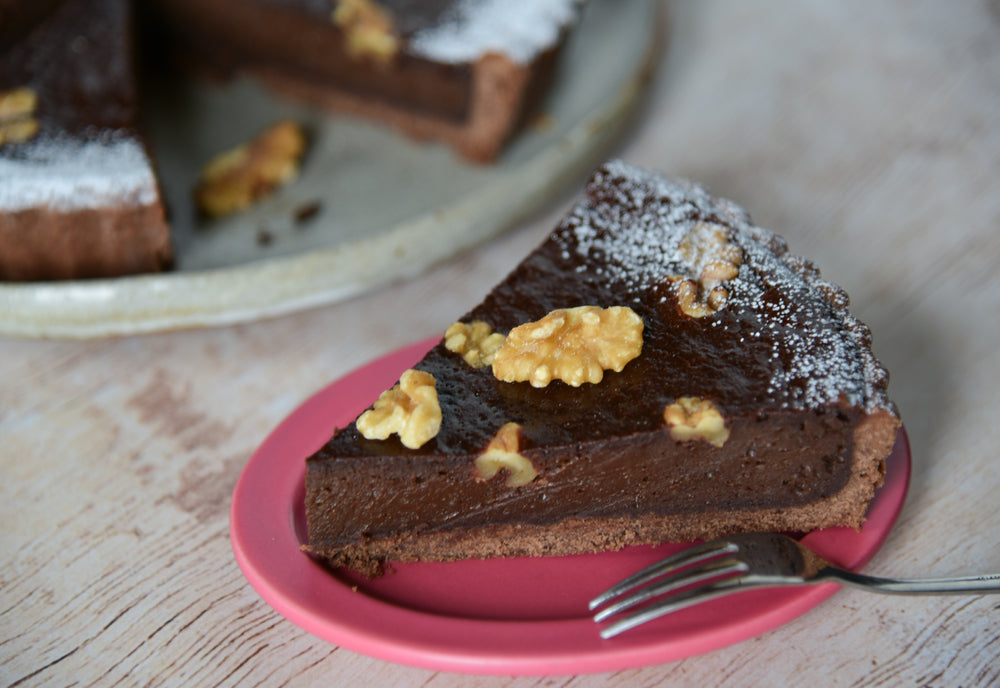 A slice of chocolate almond crust dessert topped with walnuts sits on a pink plate, with a larger cake in the background dusted with powdered sugar on a rustic wooden surface.