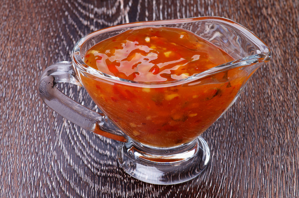 A clear glass gravy boat filled with a glossy, reddish-orange sauce sits on a dark wooden surface, showcasing a blend of ingredients including herbs and spices.