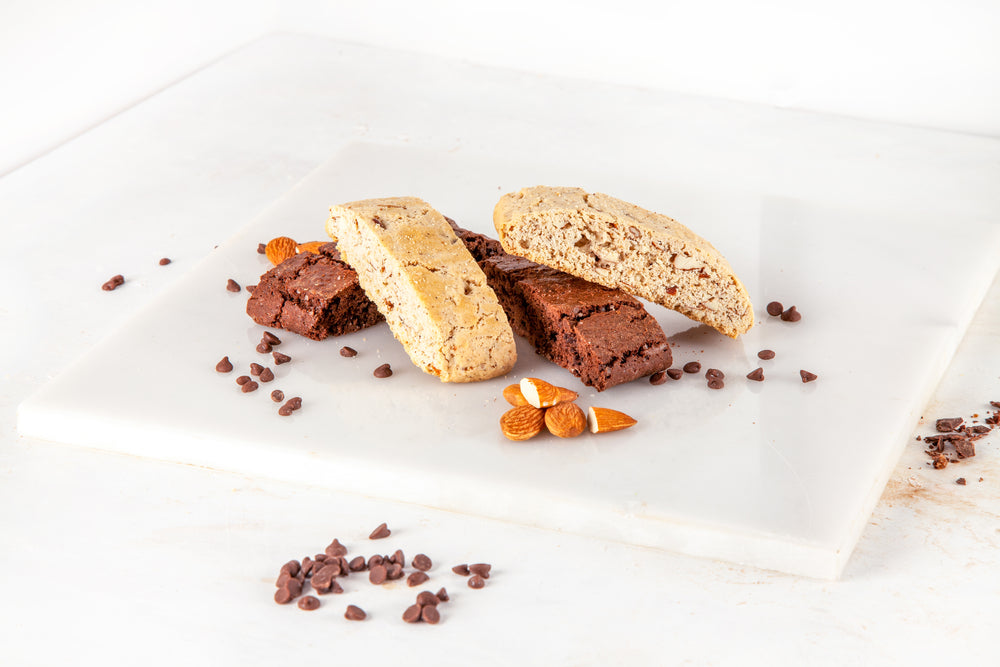 Three types of biscotti, one light and nutty and two dark chocolate, are arranged on a white marble surface, surrounded by chocolate chips and almonds.