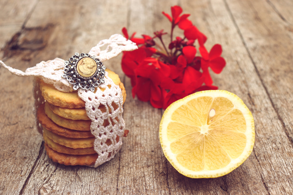 A stack of chewy lemon cookies wrapped in lace sits on a wooden surface, accompanied by a halved lemon and vibrant red flowers in the background.