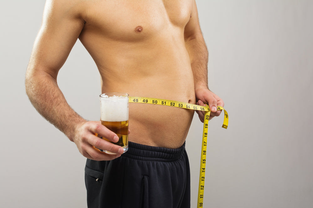 A shirtless man holds a glass of beer in one hand and measures his waist with a tape measure in the other, standing against a neutral gray background.