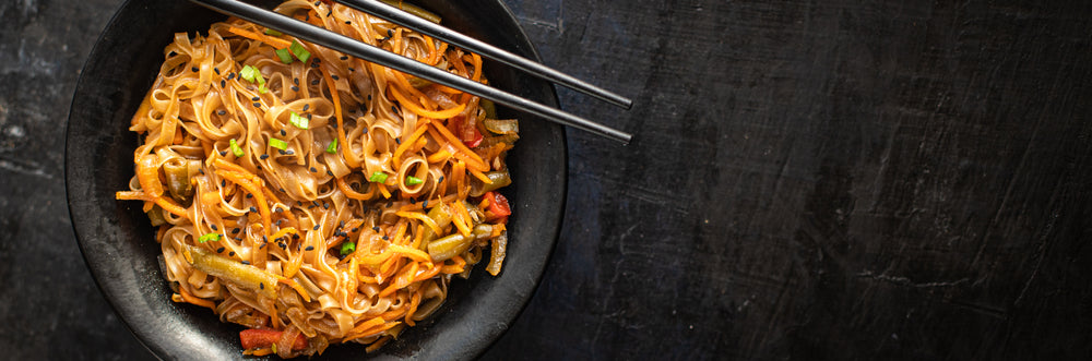 A black bowl filled with stir-fried noodles and colorful vegetables is placed on a dark surface, with a pair of chopsticks resting on the edge.