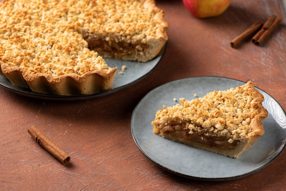 A golden-brown streusel pie sits on a dark plate, with a slice removed and displayed beside it, surrounded by cinnamon sticks and an apple on a textured surface.