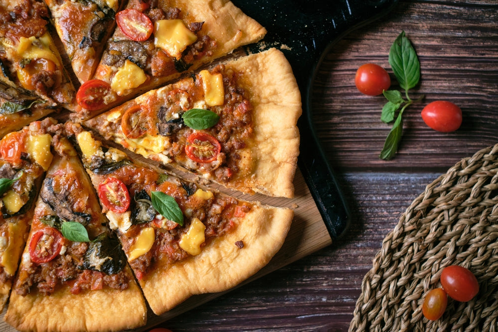 A freshly baked pizza topped with cheese, cherry tomatoes, and herbs is sliced on a wooden board, with a few whole cherry tomatoes and a woven mat nearby.