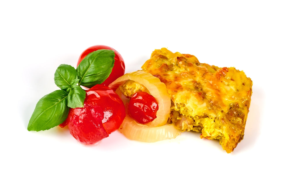 A slice of baked casserole with a golden, cheesy top is accompanied by red cherry tomatoes, a slice of onion, and fresh basil leaves on a white background.