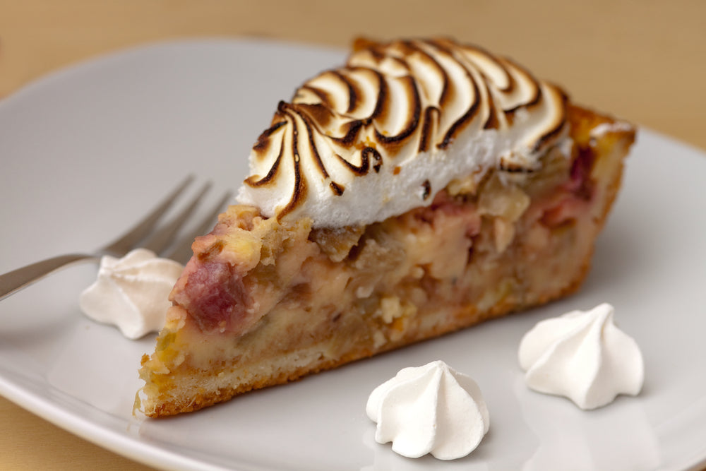 A slice of pie with a golden crust and fluffy, toasted meringue sits on a white plate, accompanied by small dollops of whipped cream and a fork resting nearby.