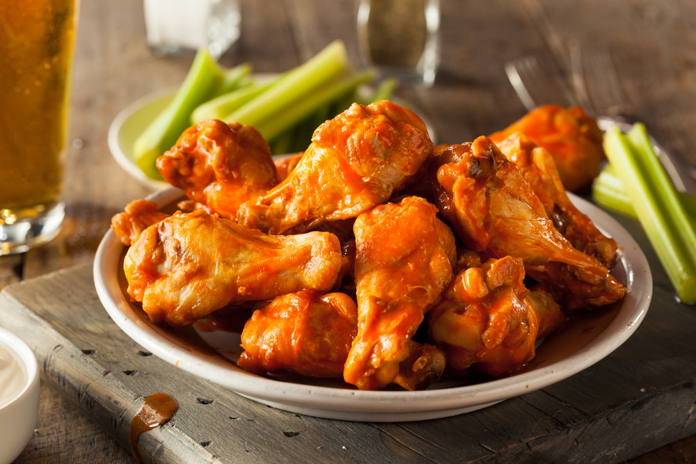 A plate of spicy buffalo chicken wings is piled high, accompanied by celery sticks and a small bowl of dipping sauce, set on a rustic wooden table with a glass of beer in the background.