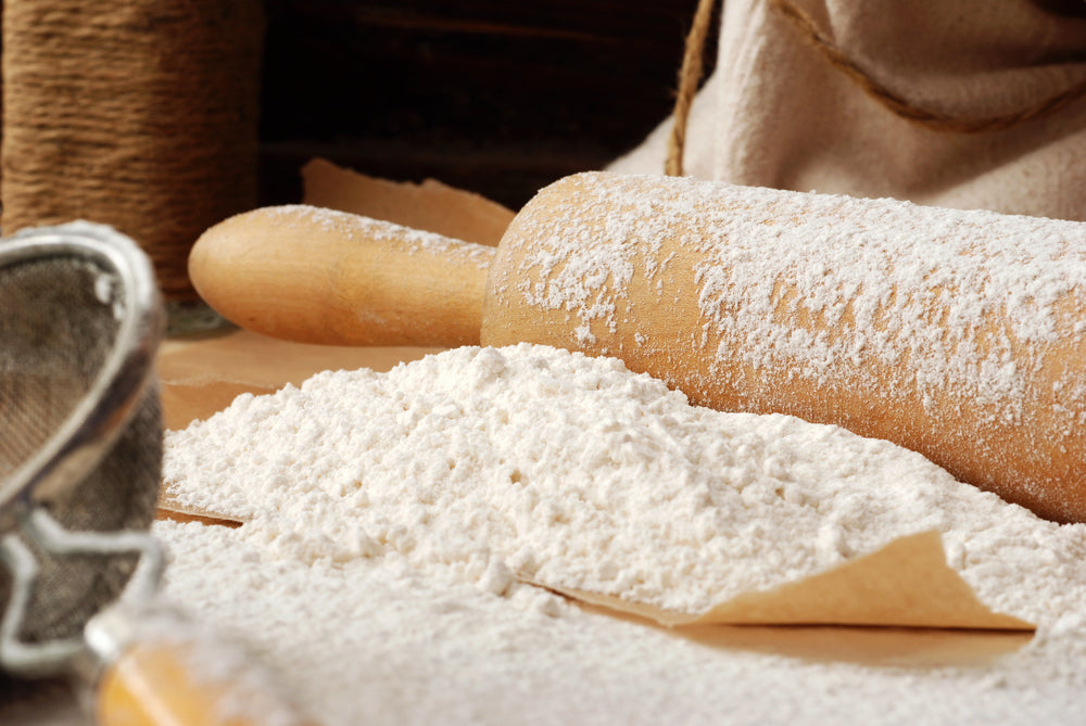 A wooden rolling pin covered in flour rests beside a pile of all-purpose low carb flour on parchment paper, with a fine mesh sieve and twine in the background, suggesting a baking setup.