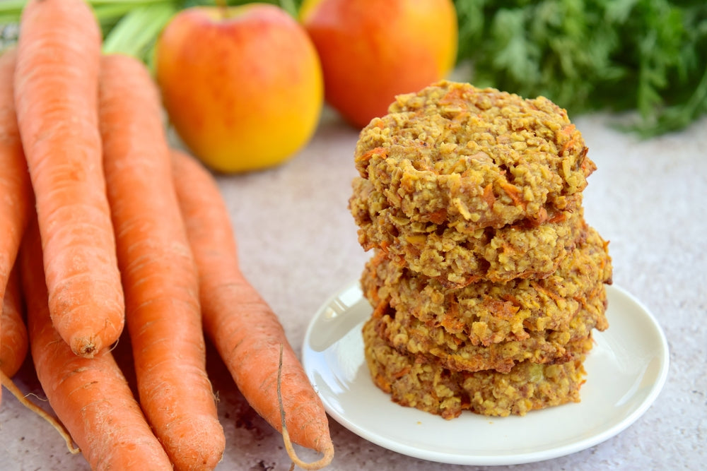 A stack of oatmeal cookies made with carrots and apples sits on a small white plate, surrounded by fresh carrots and apples on a light countertop.