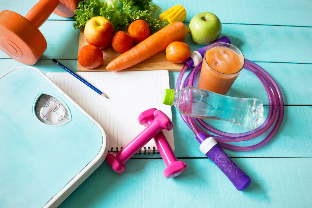 A scale, weights, a notebook with a pencil, fresh fruits and vegetables, a smoothie, a water bottle, and jump ropes are arranged on a light blue wooden surface, suggesting a healthy lifestyle.