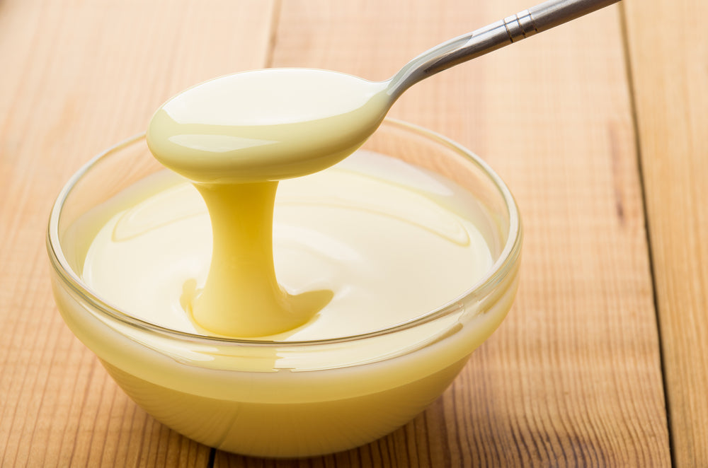 A silver spoon holds a thick, creamy yellow substance, dripping into a clear glass bowl resting on a wooden surface.