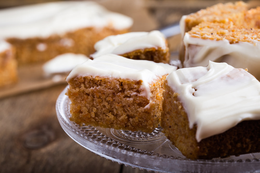 Sliced pieces of moist cake topped with creamy frosting are arranged on a decorative glass plate, with more cake in the background on a wooden surface.