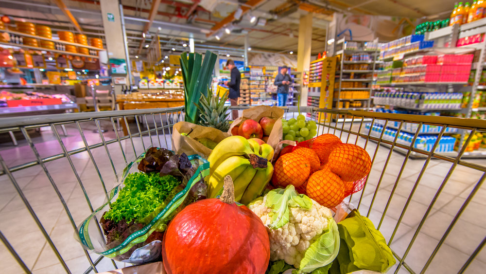 A shopping cart filled with various fresh produce, including lettuce, bananas, and oranges, is in the foreground of a vibrant grocery store with shoppers and colorful product displays in the background.