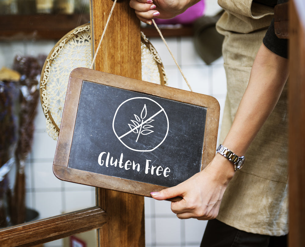 A person holds a wooden sign that reads "Gluten Free," featuring a crossed-out wheat symbol, while standing in a cozy shop with a rustic interior.