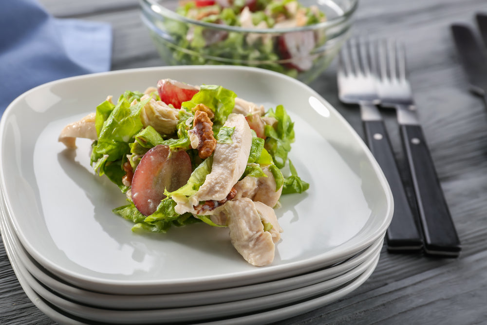 A plate of salad featuring lettuce, chicken, walnuts, and tomatoes is set on a wooden table, with a bowl of mixed salad and utensils in the background.