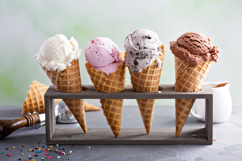 Four ice cream cones with different flavors—vanilla, strawberry, cookies and cream, and chocolate—are displayed in a wooden holder on a gray surface, with sprinkles and a small pitcher nearby.