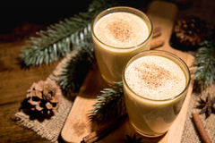 Two glasses of creamy, egg-free low carb eggnog are garnished with a sprinkle of nutmeg, placed on a wooden board surrounded by pine branches and pinecones, evoking a cozy holiday atmosphere.