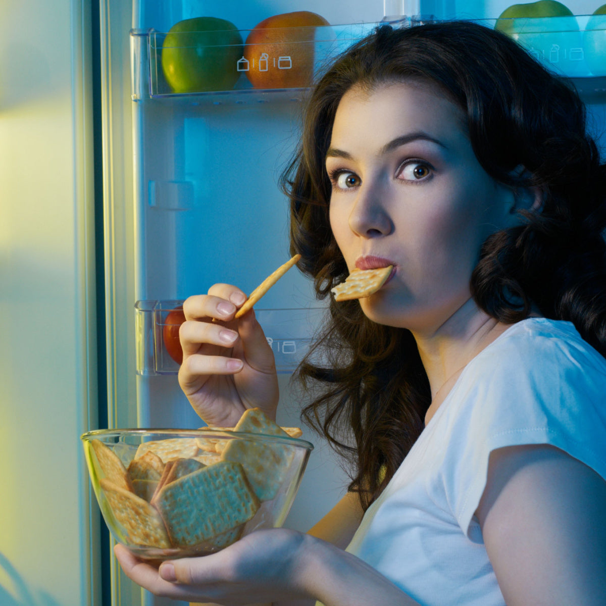 A woman with long, dark hair stands in front of an open refrigerator, holding a bowl of crackers in one hand and a cracker in her mouth, looking surprised or amused.
