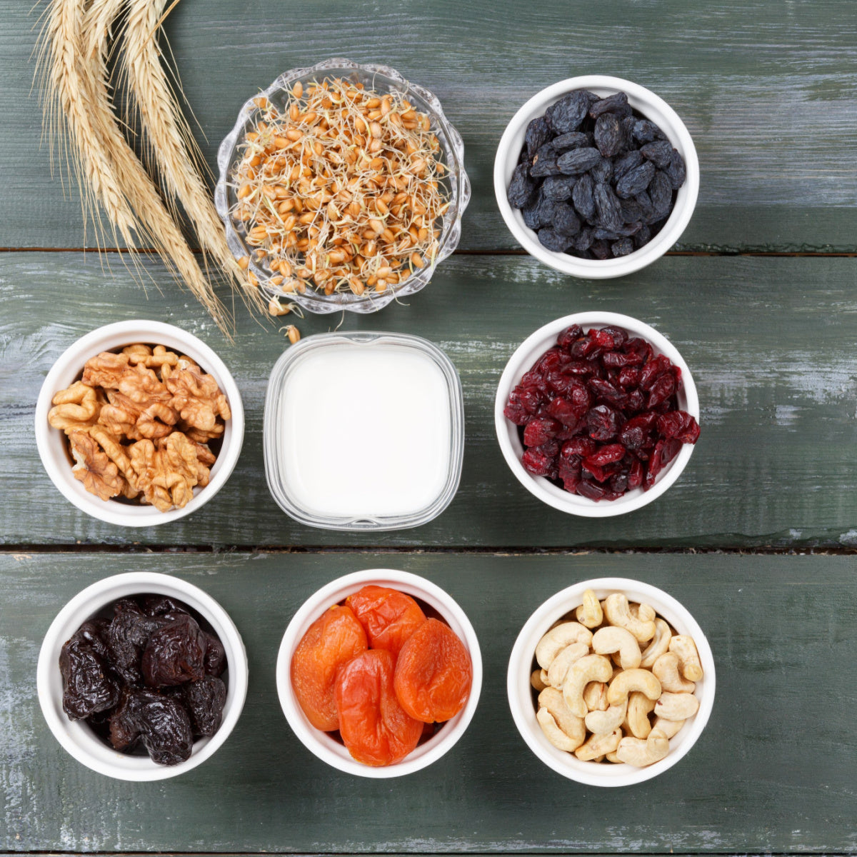 A variety of nuts, dried fruits, and sprouts are arranged in white bowls on a rustic wooden table, with a small container of milk in the center, showcasing healthy snack options.