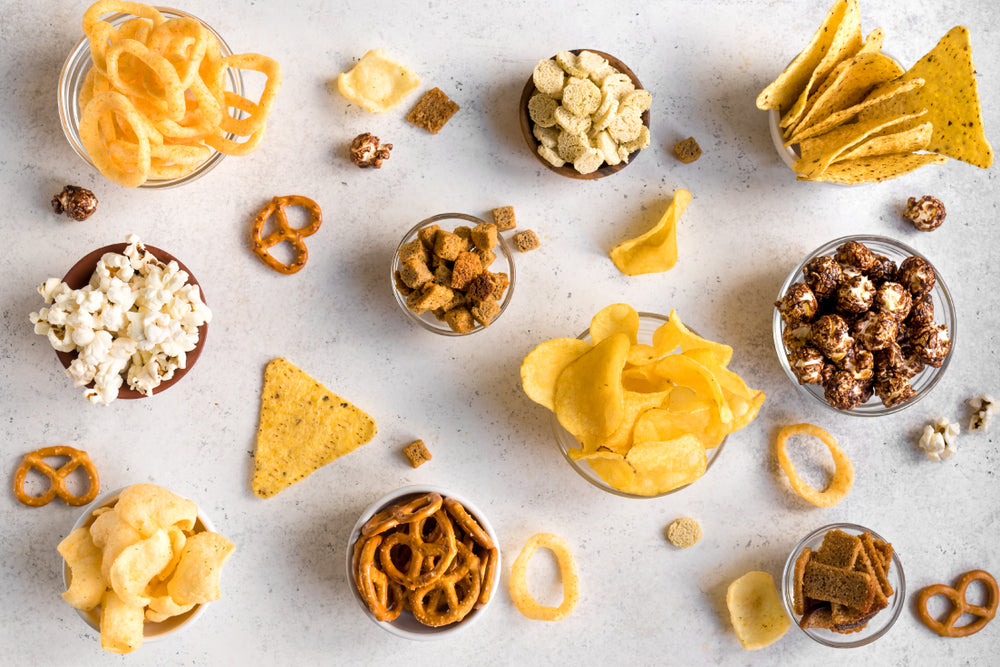 A variety of low-carb snacks, including popcorn, chips, pretzels, and crackers, are displayed in clear bowls on a light surface, showcasing options for keto dieters.