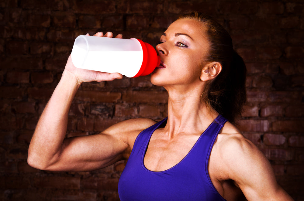 A fit woman in a purple tank top drinks from a red and white shaker bottle while standing in front of a brick wall, suggesting a gym or workout setting.
