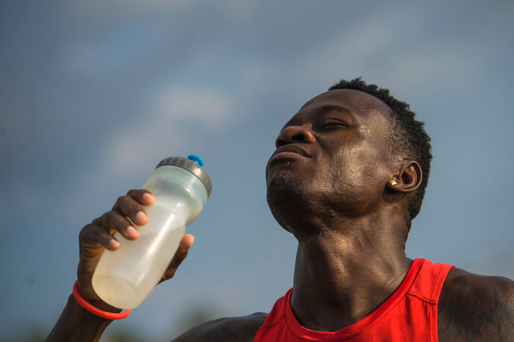 A man in a red tank top drinks from a sports bottle, tilting his head back with a focused expression against a blue sky, emphasizing hydration for effective sports recovery.