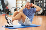 A smiling older man performs crunches on a blue exercise mat in a gym, surrounded by fitness equipment, showing enthusiasm for his workout.