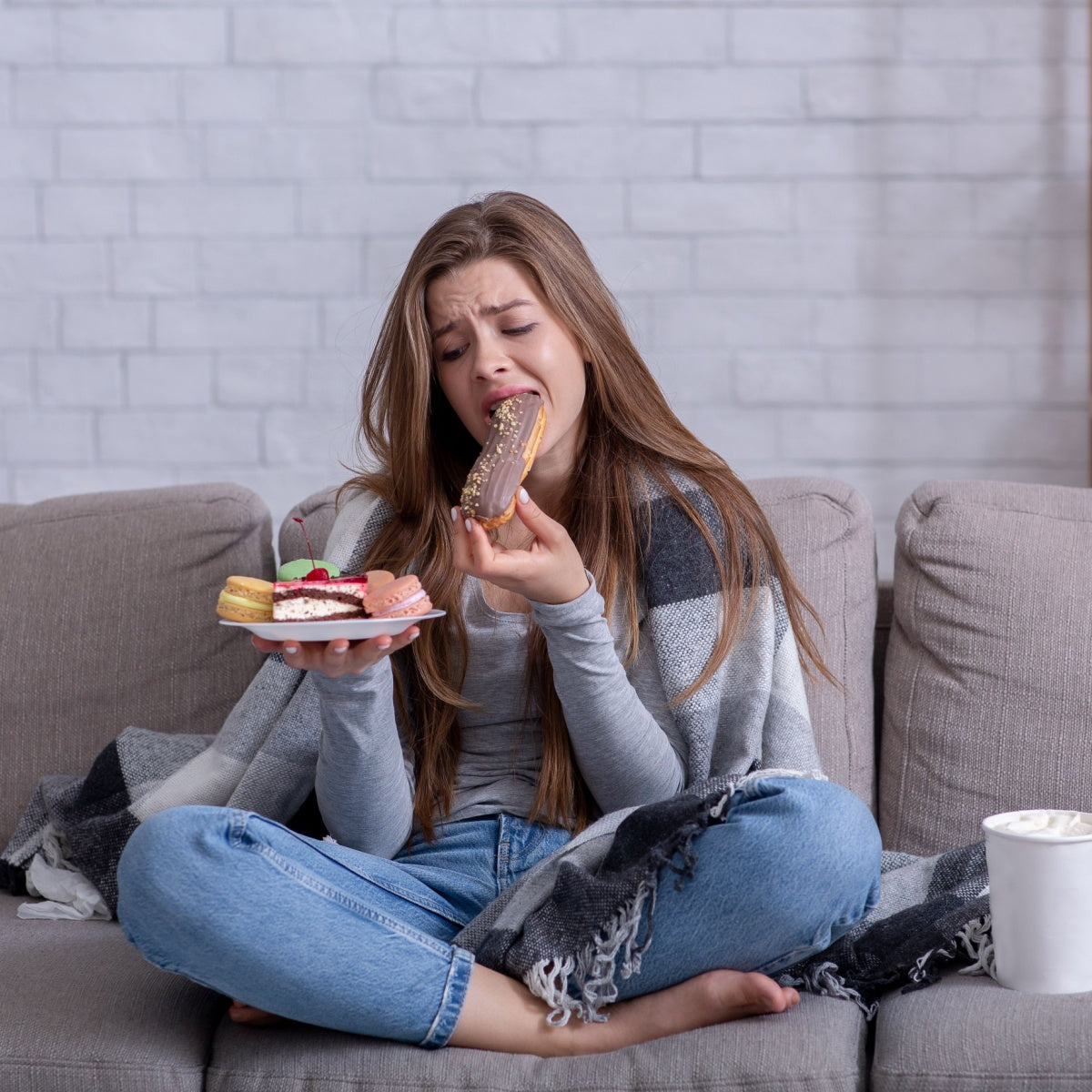 A young woman sitting cross-legged on a couch, wearing a gray top and wrapped in a blanket, looks overwhelmed while eating a chocolate-covered pastry from a plate of assorted desserts.