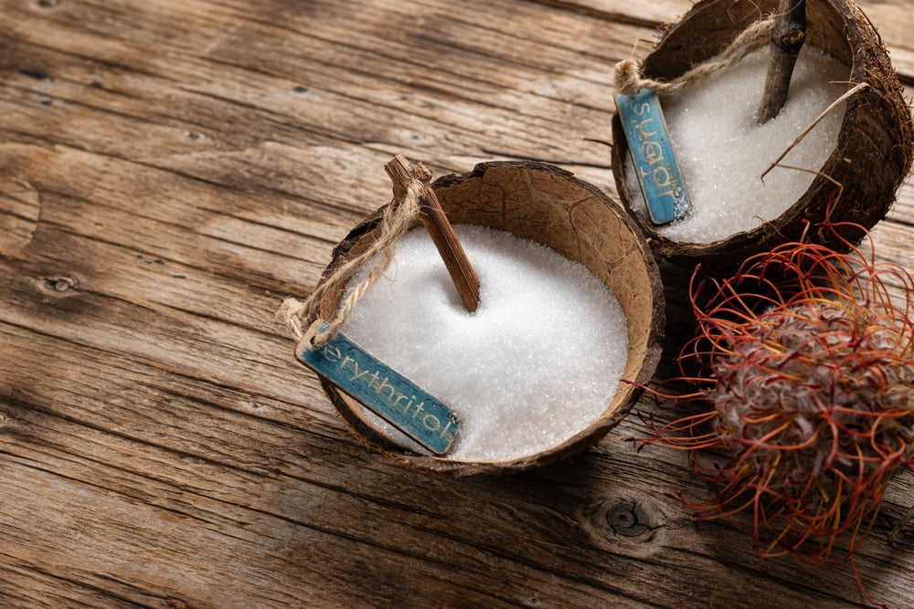 Two coconut bowls filled with white granulated sweeteners labeled "erythritol" and "sugar" sit on a rustic wooden surface, accompanied by a spiky red fruit nearby.